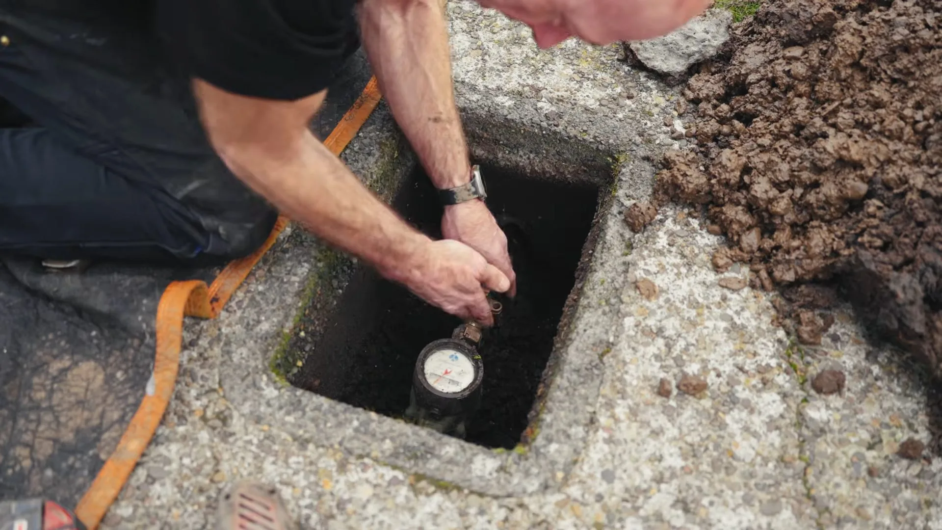 Man kneeling beside an open in-ground box and reaching down toward the water meter inside