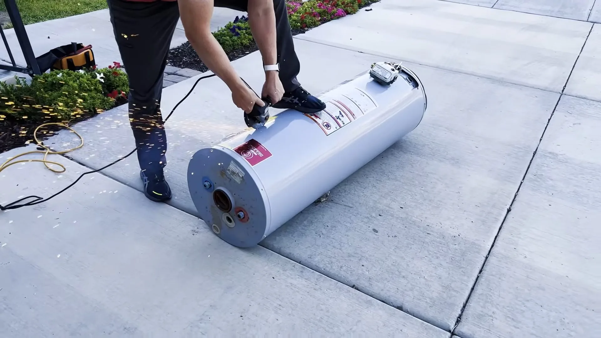 Two people tilting and rolling a white water heater tank across a concrete patio