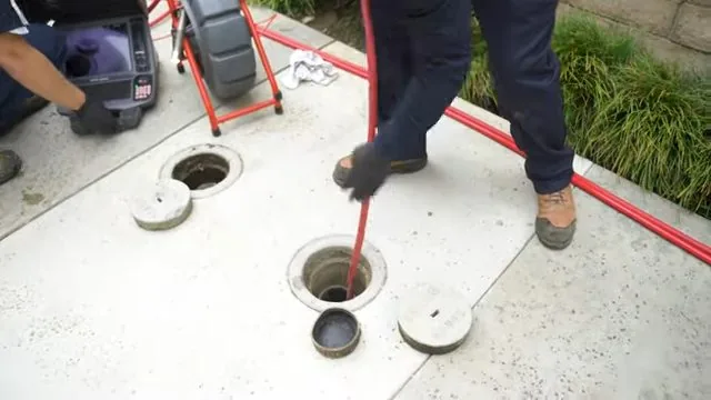 Man standing on a concrete pad feeding a red drain cable into a round floor drain, with a camera case and reel behind him