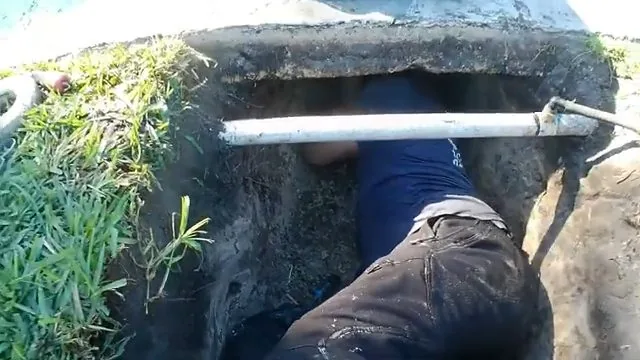 Worker in an excavated pit with a white pipe crossing over him and grass at the edge of the hole