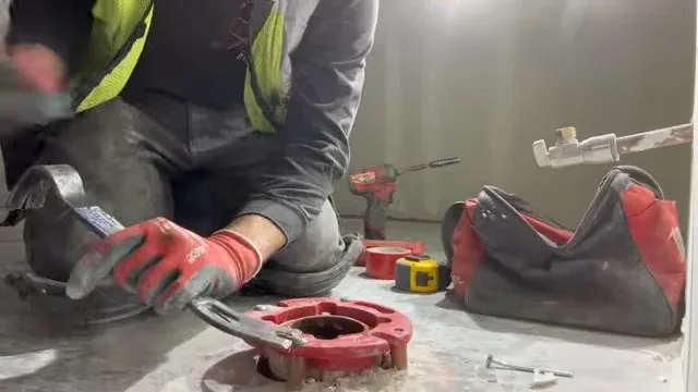 Worker in a high-visibility vest kneeling beside a floor flange with a red wax ring and wrenches, and a tool bag nearby