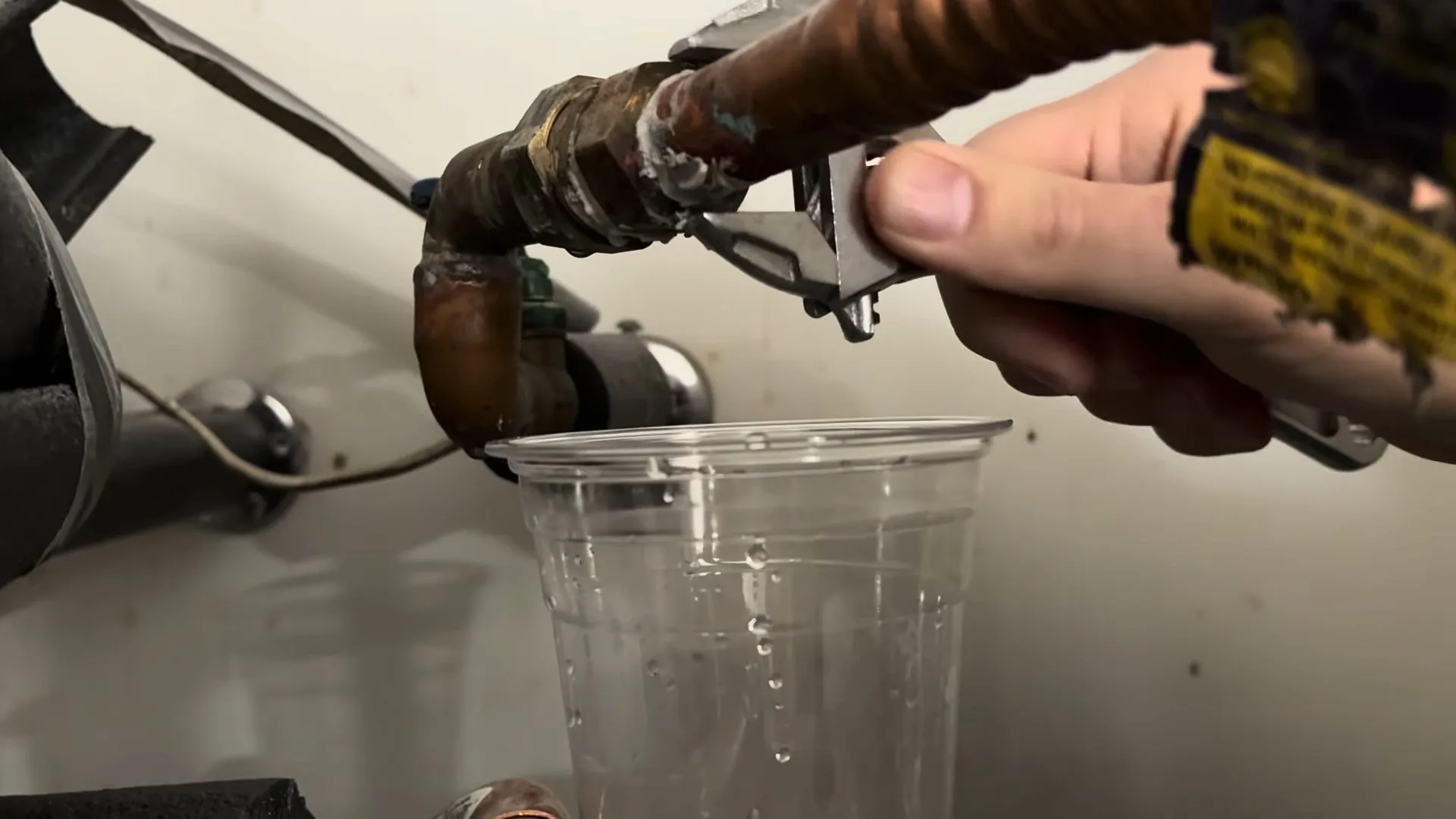 Hand holding a small adjustable wrench on a copper fitting above a clear cup, with a gray water heater behind